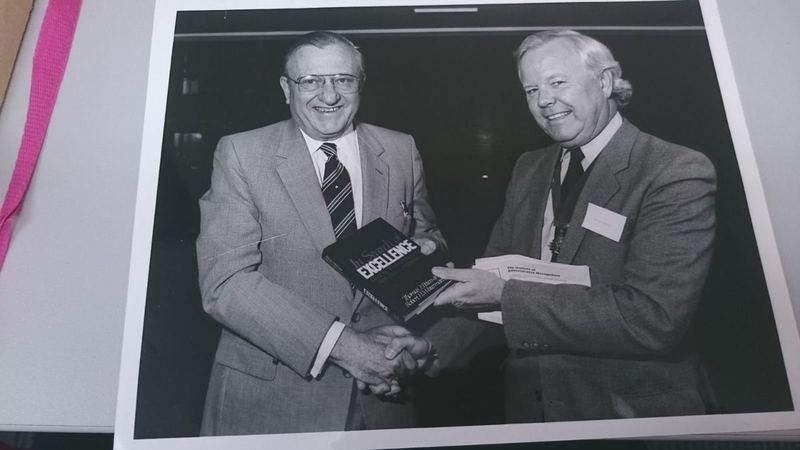 Archive photograph of two men shaking hands while presenting a copy of the book In Search of Excellence, with Institute of Administrative Management papers visible